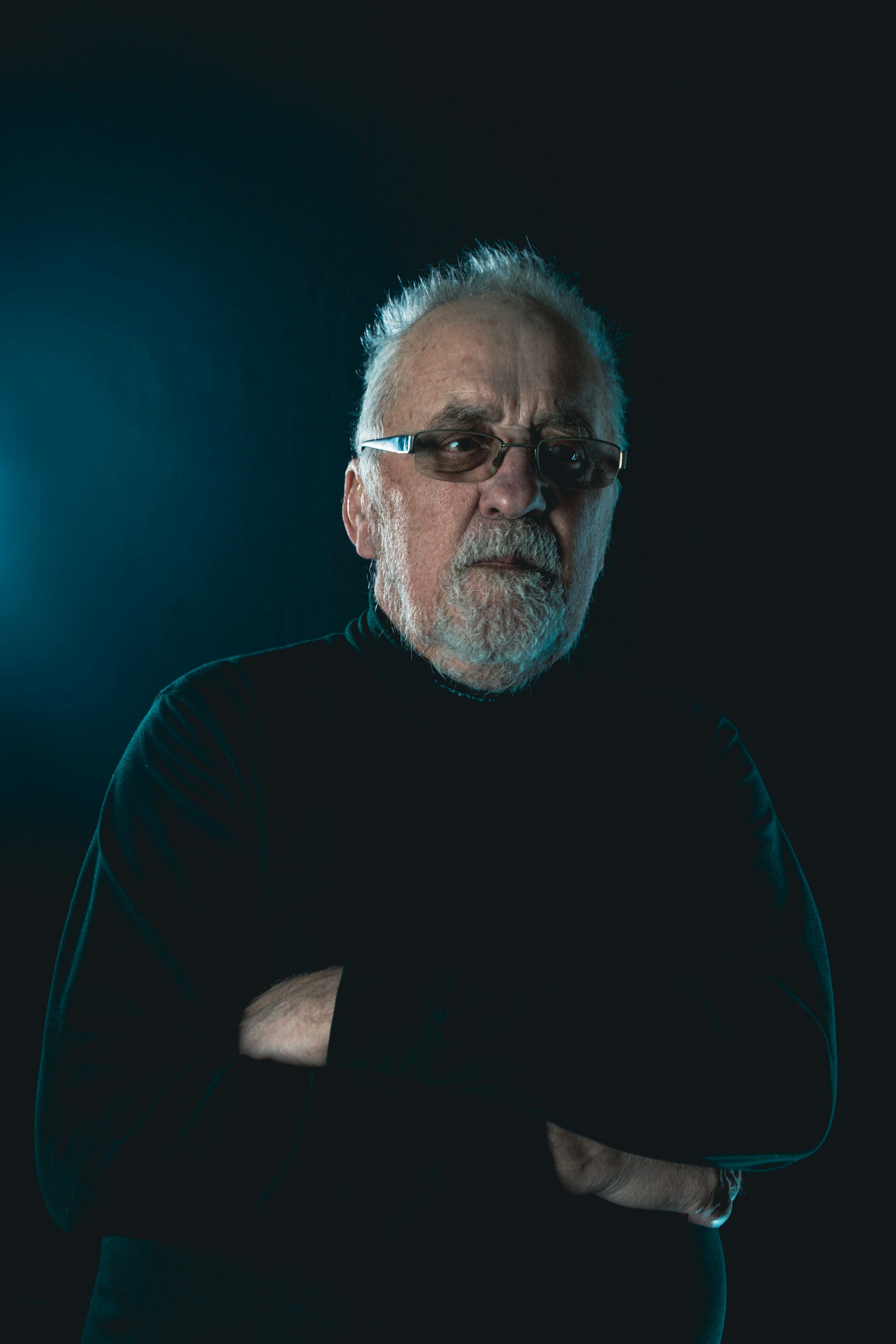 Portrait: an older man with white hair and a beard, arms folded, against a dark teal background