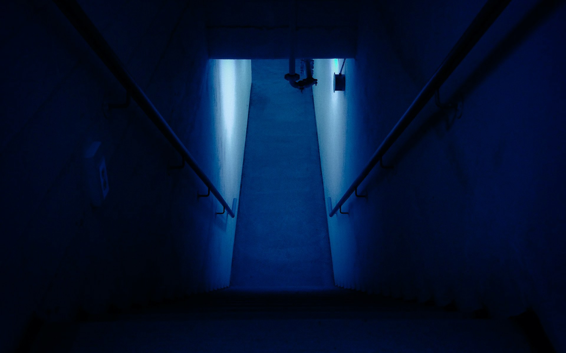 A bare concrete institutional interior: an open stair rising on the left, a long corridor lit by ceiling fittings, glass doors at the far end